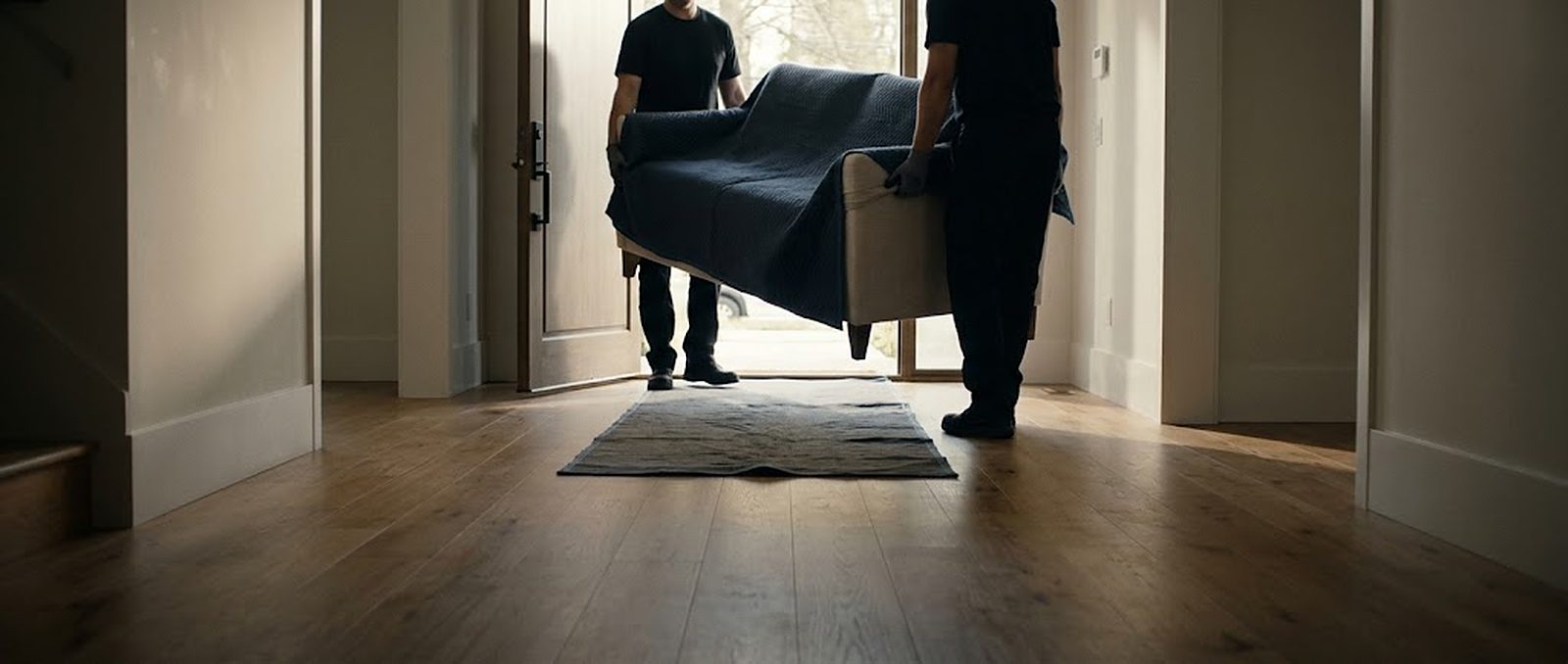 Two crew members carrying a blanket-wrapped armchair through a doorway into a finished room