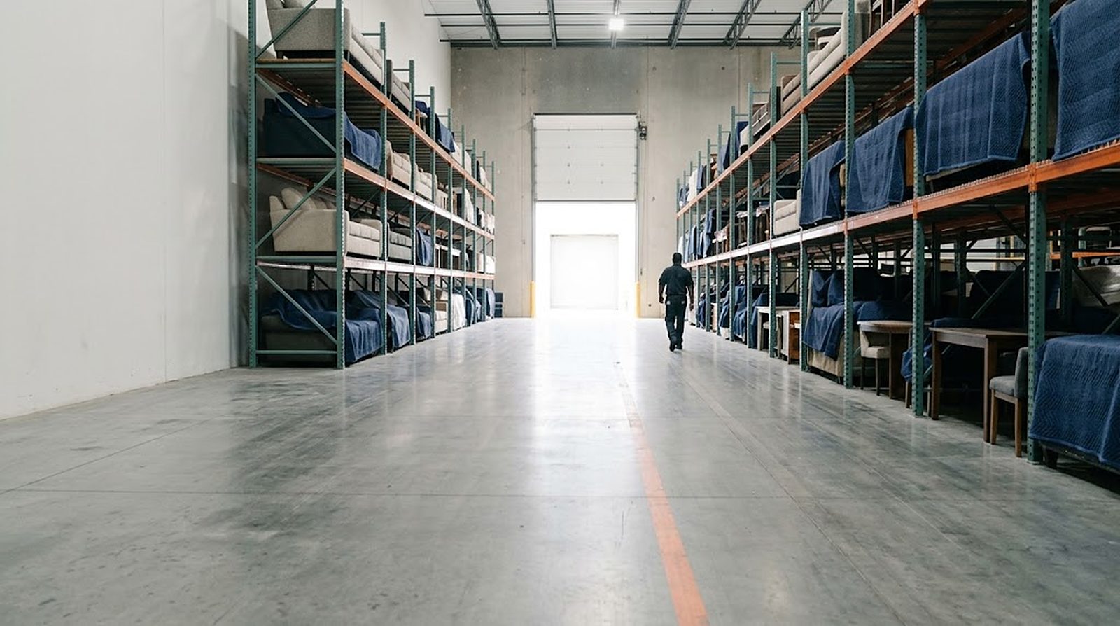 A warehouse aisle lined with racked, blanket-wrapped furniture, a member of the team walking toward the open dock door
