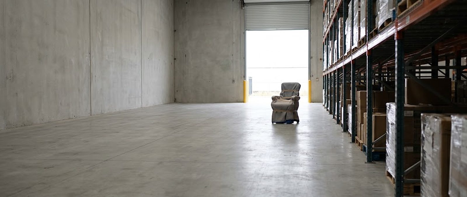 A quiet warehouse aisle looking toward the open dock door, a single wrapped piece waiting